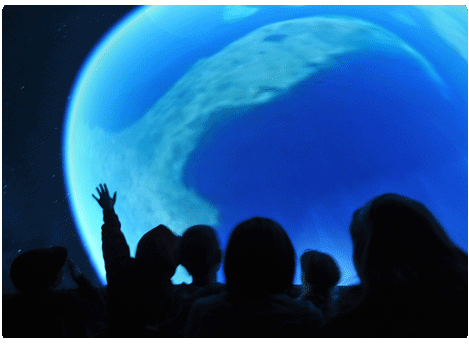 Audience members silhouetted in a planetarium dome, looking up at a projected blue Earth